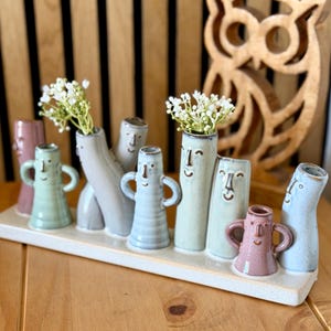 May include: A ceramic display featuring multiple small vases in various pastel colours, each shaped with a simple face and some with side handles. Two vases contain small white flowers. The vases are arranged on a rectangular white base.