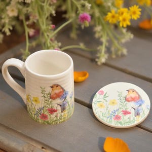 May include: A white ceramic mug and coaster set. The mug has a curved handle and is decorated with a colourful illustration of a robin bird, wildflowers, and green grass. The coaster has a matching design. The set is on a grey wooden surface.