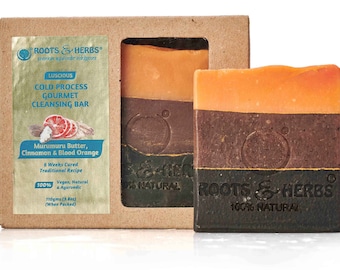 Könnte beinhalten: Zwei handgemachte Seifenstücke in einem Karton. Die Seife ist in Braun, Orange und Schwarz geschichtet. Das Etikett auf der Schachtel lautet "Roots & Herbs" und "Luscious Cold Process Gourmet Cleansing Bar". Die Seife wird aus Murumuru-Butter, Zimt und Blutorange hergestellt. Sie ist 100% vegan, natürlich und ayurvedisch.
