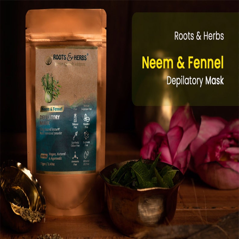 May include: A brown pouch with a label that reads "Roots & Herbs Neem & Fennel Depilatory Mask". The pouch contains a hair removal powder. The pouch is sitting on a wooden surface with a bowl of green leaves and a pink flower.