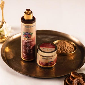 Może przedstawiać: Cylindryczna butelka Roots & Herbs Flawless Skin Activator oraz mały słoiczek Flawless Skin Ubtan, oba z ilustracjami fig i złotymi nakrętkami. Produkty umieszczono na okrągłej mosiężnej tacy wraz z małą miseczką z brązowym proszkiem.
