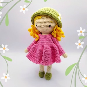 Könnte beinhalten: Eine gehäkelte Puppe mit blonden Haaren, die ein rosa Kleid und einen grünen Hut trägt.