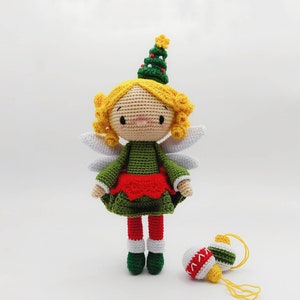 Può includere: Una bambola di angelo di Natale all'uncinetto con capelli biondi, un vestito verde, una gonna rossa e ali bianche. La bambola tiene un piccolo ornamento natalizio rosso e bianco.