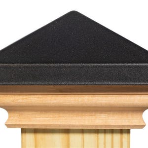 May include: A black, triangular post cap sits atop a wooden post. The cap has a textured surface and a slightly rounded edge. The wooden post has a decorative, carved design.