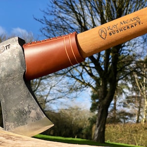May include: A Ray Mears Bushcraft axe with a brown leather handle and a wooden shaft. The axe is resting on a piece of wood with a green grassy background.