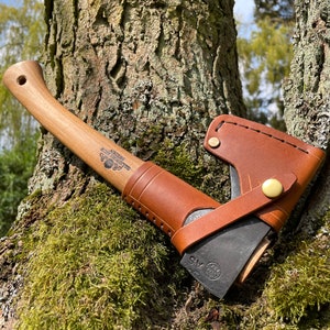 May include: A brown leather sheath with a small axe inside. The axe has a wooden handle and a black metal head. The sheath has a brass button and stitching. The axe is resting against a tree trunk.