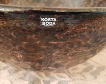 May include: A dark brown glass bowl with a textured surface. The bowl has a label that reads "Kosta Boda Sweden Glass".