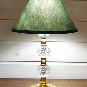 Op de afbeelding: Een vintage tafellamp met een groene lampenkap van stof en een kristallen voet. De voet heeft twee heldere kristallen bollen en een gouden metalen voet.