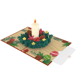 May include: A 3D pop-up card featuring a white candle in a green wreath with red berries, pine cones, and gold stars. The card has a wooden background with a small envelope attached.