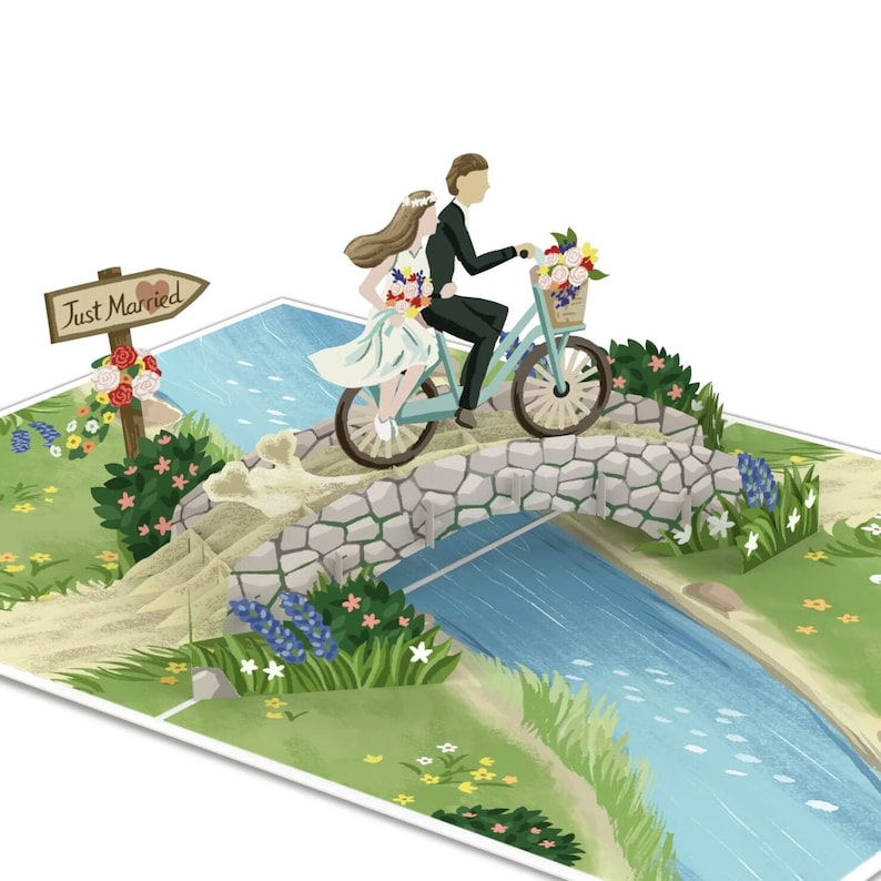 May include: A pop-up card featuring a bride and groom riding a blue bicycle over a stone bridge. The bride is wearing a white dress and a flower crown. The groom is wearing a black suit. The card has a sign that says "Just Married" with a heart.