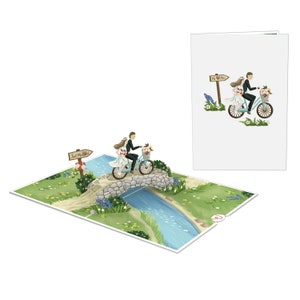 May include: A 3D pop-up card featuring a bride and groom riding a bicycle over a stone bridge. The card is open and the pop-up is extended. The bride and groom are wearing white and black clothing, respectively. The bicycle is blue and has a basket of flowers on the handlebars. The bridge is made of stone and has a small stream flowing underneath it. The card is set against a green grassy background with blue flowers.