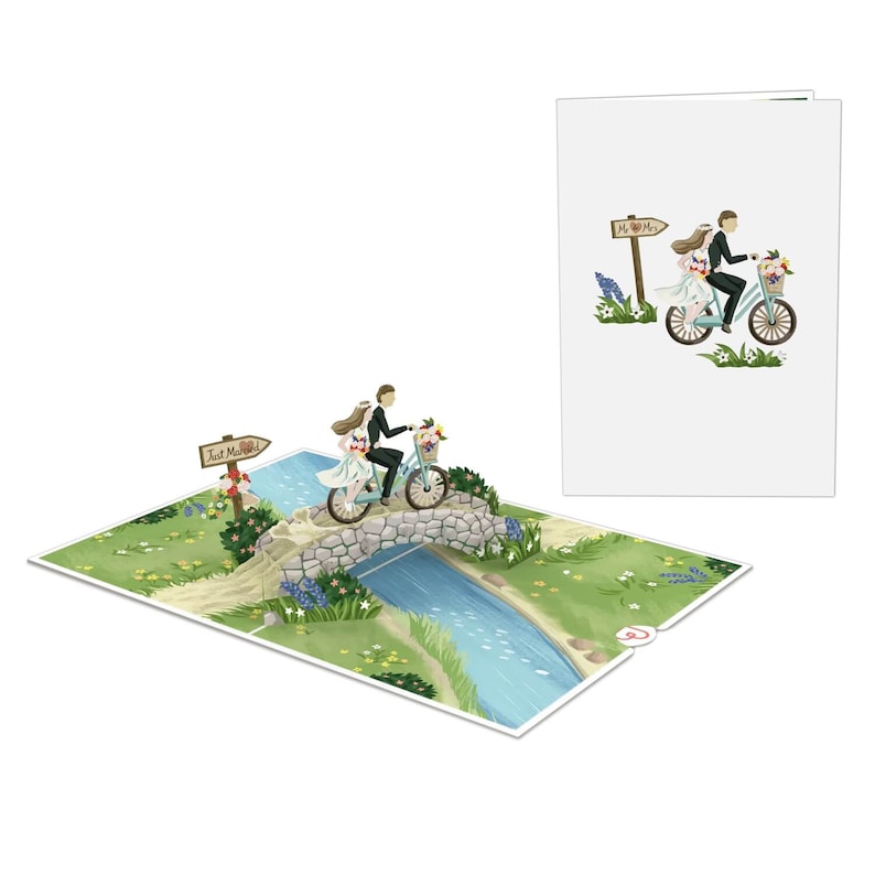 May include: A 3D pop-up card featuring a bride and groom riding a bicycle over a stone bridge. The card is open and the pop-up is extended. The bride and groom are wearing white and black clothing, respectively. The bicycle is blue and has a basket of flowers on the handlebars. The bridge is made of stone and has a small stream flowing underneath it. The card is set against a green grassy background with blue flowers.