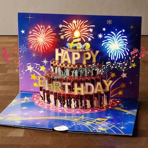 Peut inclure: Une carte d'anniversaire pop-up avec un design de gâteau au chocolat et l'inscription dorée "Happy Birthday". La carte a un fond bleu avec des feux d'artifice et des notes de musique. Une bougie allumée est posée sur le gâteau.
