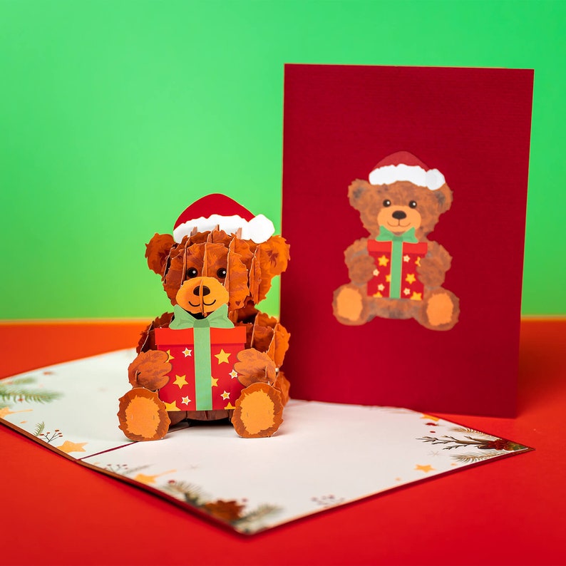 May include: A 3D pop-up card featuring a brown teddy bear wearing a red Santa hat and holding a red and green gift box. The card is open and the bear is standing out from the card. The background is a red surface.