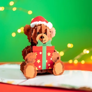 May include: A brown teddy bear wearing a red Santa hat holds a red and green gift box with white stars. The bear is sitting on a white card with a red background.