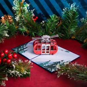 May include: A red and white 3D pop-up card featuring a cable car with a snowy mountain scene in the background. The card is open and the cable car is raised.