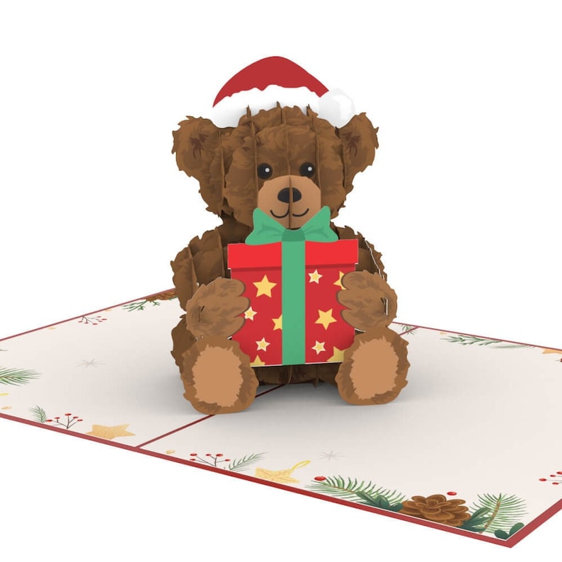 May include: A brown teddy bear wearing a red Santa hat holds a red and green gift box with gold stars. The bear is sitting on a white card with a wintery border.