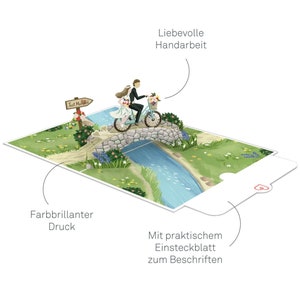 May include: A 3D pop-up card featuring a bride and groom riding a bicycle over a stone bridge. The card has a 'Just Married' sign and a blue river. The card is handmade and has a blank insert for writing a message.