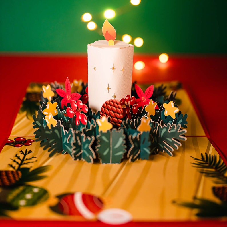 May include: A white candle with a red flame sits in the center of a paper pop-up card. The candle is surrounded by a green wreath with red berries, yellow stars, and pine cones. The card is on a red surface.