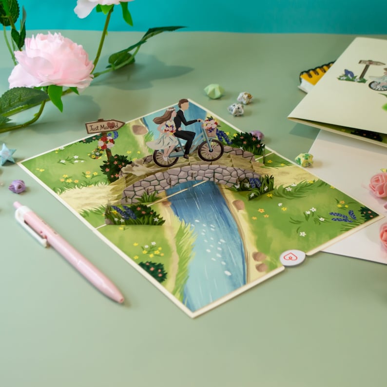 May include: A pop-up card featuring a couple riding a bicycle over a stone bridge. The card is decorated with flowers and a sign that says "Just Married".