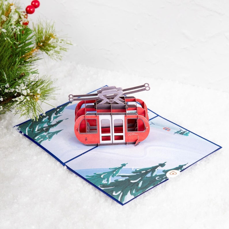 May include: A red and white 3D pop-up card featuring a cable car with a snowy mountain scene in the background.