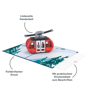 May include: A red and white 3D pop-up card featuring a cable car in a snowy mountain scene. The card has a blank insert for writing a message. The card is handmade and features a vibrant color print.