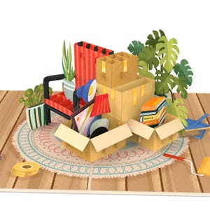 May include: A pop-up card depicting moving day. The card features cardboard boxes, furniture, plants, and a rug. A red and black chair, a blue lamp, and a stack of books are visible. The scene is set on a wooden floor.