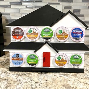 May include: A house-shaped coffee pod holder in white and black, with a red door. It displays various coffee and hot chocolate pods, including brands like McCafe, Green Mountain, and Swiss Miss. The holder is made of wood.