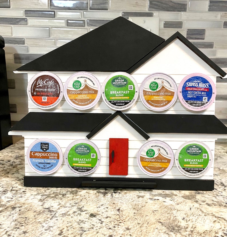 May include: A house-shaped coffee pod holder in white and black, with a red door. It displays various coffee and hot chocolate pods, including brands like McCafe, Green Mountain, and Swiss Miss. The holder is made of wood.
