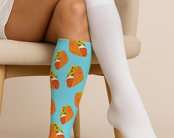 May include: A person seated on a chair, wearing a pair of knee-high socks. One sock is solid white, while the other is light blue with a repeating pattern of orange tacos with green and red fillings. The socks are made of a soft, stretchy material.