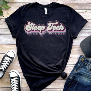 Op de afbeelding: Zwart T-shirt met een retro-grafische afbeelding met de tekst "Sleep Tech" in kleurrijke, golvende letters.