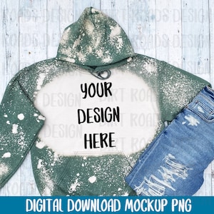 Könnte beinhalten: Grüner, gebleichter Hoodie mit einem weißen Designbereich, auf dem "YOUR DESIGN HERE" steht. Der Hoodie liegt auf einem weißen Holzgrund mit einer blauen Jeans.