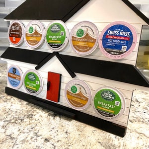 May include: A house-shaped coffee pod holder made of wood, painted white with a black roof and trim, and a red door. It holds various coffee and hot chocolate pods, including McCafe, Green Mountain, and Swiss Miss.