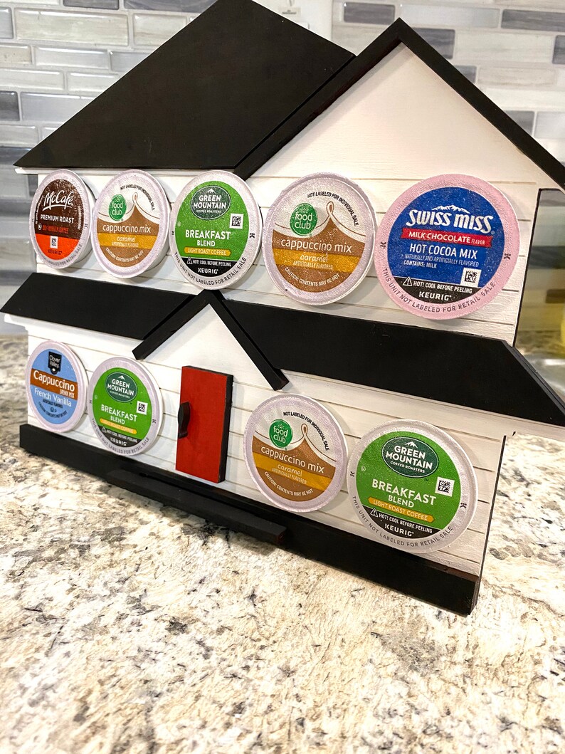 May include: A house-shaped coffee pod holder made of wood, painted white with a black roof and trim, and a red door. It holds various coffee and hot chocolate pods, including McCafe, Green Mountain, and Swiss Miss.