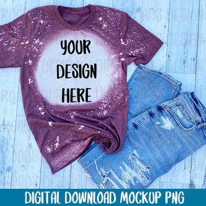 May include: A maroon t-shirt with a white bleach design. The design is a circle with the text "YOUR DESIGN HERE" in black. The t-shirt is laying on a light wood background with a pair of blue jeans.