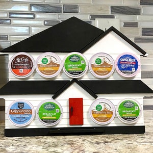 May include: A house-shaped coffee pod holder in white and black, with a red door. It displays various coffee and hot chocolate pods, including McCafe, Food Club, Green Mountain, and Swiss Miss brands. The holder is designed to store coffee pods.