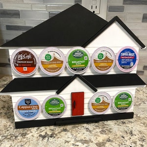 May include: A white, house-shaped wooden K-cup holder with a black roof and base. It displays various coffee and hot chocolate pods, including McCafe, Green Mountain, and Swiss Miss. The holder has a red door.