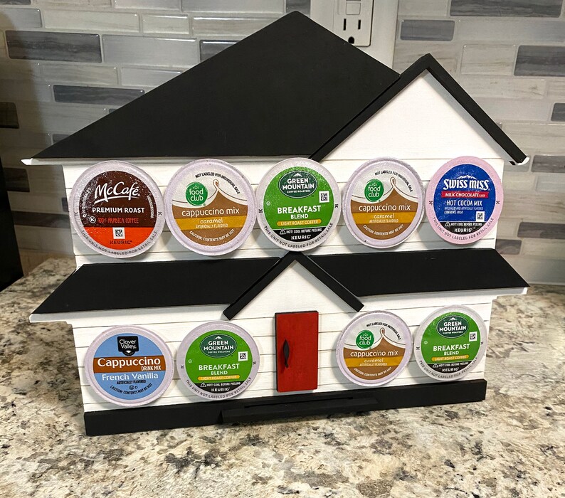 May include: A white, house-shaped wooden K-cup holder with a black roof and base. It displays various coffee and hot chocolate pods, including McCafe, Green Mountain, and Swiss Miss. The holder has a red door.