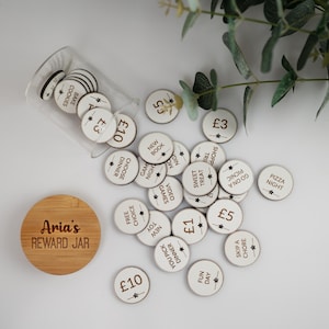 May include: A wooden reward jar with a white lid and a label that says "Aria's Reward Jar". The jar is filled with white wooden tokens with different amounts of money and activities written on them, such as "£10", "£5", "£3", "£1", "Free Choice", "Game Night", "Sweet Treat", "Pizza Night", "You Pick Dinner", "Fun Day", "Skip a Chore", "Go on a Picnic", "New Book", "New Toy", "Bake Cookies", and "Choose Dinner".