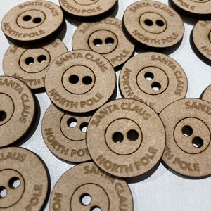 May include: A close-up of a pile of wooden buttons with the text "Santa Claus North Pole" printed on them. The buttons are brown and have two holes each.