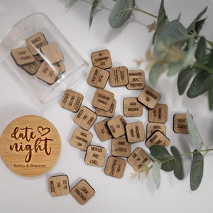 May include: A wooden jar with a lid that says "date night" with the names Bobby & Shannon. The jar is filled with small wooden squares with date night ideas written on them. The jar is sitting on a white surface with a green plant in the background.