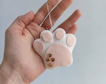 May include: A felt paw print ornament in white and light pink, with gold sequins. The paw print has a scalloped edge and a white string for hanging. A charming, handmade decoration.