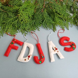 May include: Felt Christmas ornaments spelling out "F D J A S". The letters are red and white, decorated with sequins. Green evergreen branches are at the top, with a light blue background. Each letter has a string for hanging.