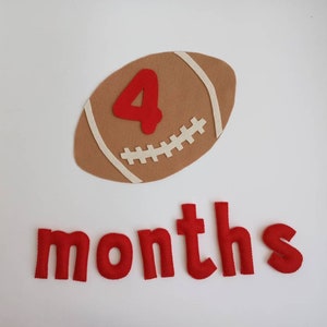 Può includere: Un pallone da football americano in feltro marrone con il numero 4 in rosso e la parola "months" in lettere rosse. Il pallone ha cuciture e strisce bianche. Lo sfondo è bianco.