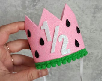May include: A pink glitter crown with a watermelon design. The crown has black seed accents and the number "1/2" in white. A green pom-pom trim lines the bottom edge. White elastic straps are attached for wear.