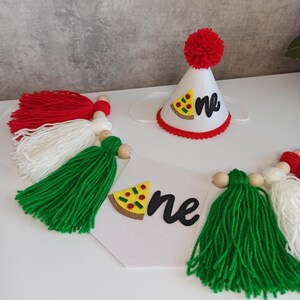 May include: A white glittery party hat with a red pom-pom, a pizza slice cutout, and the word one in black script, paired with a banner featuring red, white, and green yarn tassels, wooden beads, and a white pennant with a pizza slice and one.