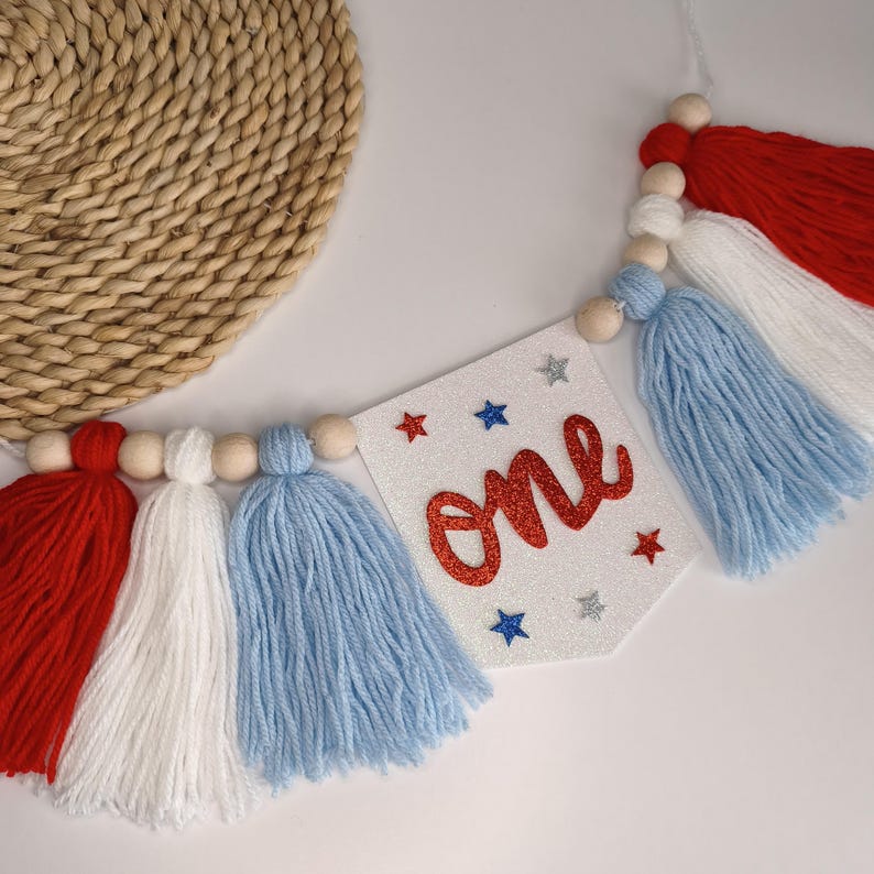 May include: A decorative banner featuring a white glittery pennant with the word one in red glitter script, surrounded by small red, blue, and silver glitter stars. The banner is strung with natural wooden beads and red, white, and blue yarn tassels.