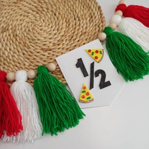 May include: A festive banner with the number "1/2" in black glitter, adorned with pizza slice accents. The banner is part of a garland featuring red, white, and green tassels, wooden beads, and a woven straw background.