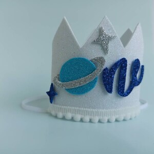 Puede incluir: Corona de fieltro blanco con purpurina azul y plateada, un planeta azul con un anillo plateado, una estrella azul y la palabra "one" en purpurina azul.