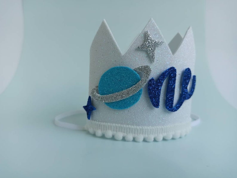 Puede incluir: Corona de fieltro blanco con purpurina azul y plateada, un planeta azul con un anillo plateado, una estrella azul y la palabra "one" en purpurina azul.